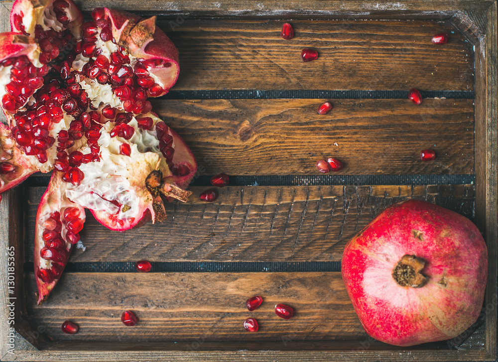 Fresh ripe seasonal pomegranates, one cut in pieces in rustic wooden tray, top view, copy space, hor