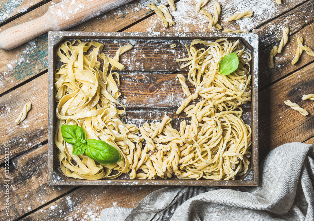 Various homemade fresh uncooked Italian pasta with flour and green basil leaves in wooden tray over 