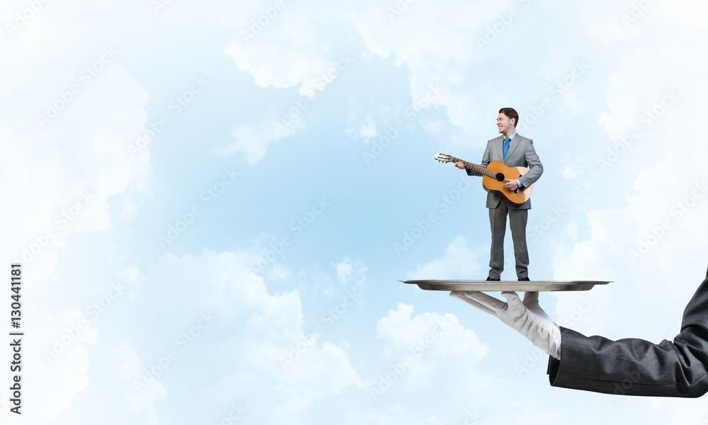 Businessman on metal tray playing acoustic guitar against blue sky background