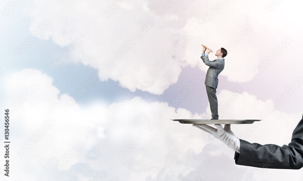 Businessman on metal tray playing fife against blue sky background