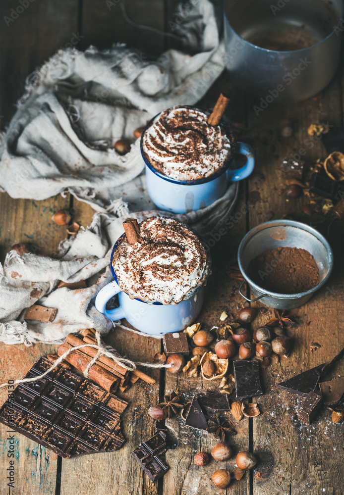 Hot chocolate in mugs with whipped cream and cinnamon, spices, nuts and cocoa powder on rustic woode