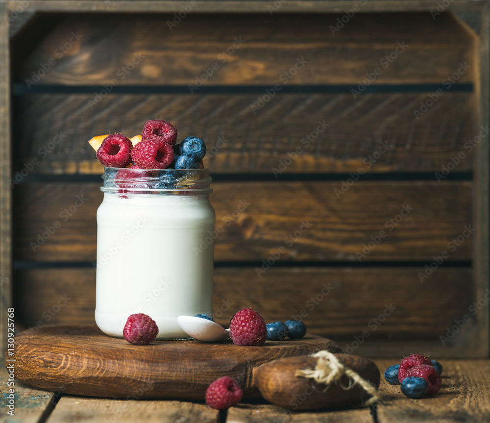 Glass jar of white yogurt with fresh garden berries and peach on serving board over rustic table, wo