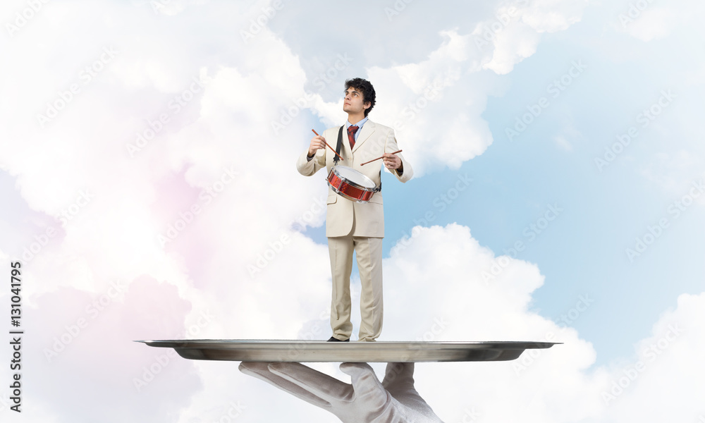 Young businessman on metal tray playing drums against blue sky background