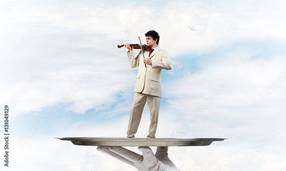 Businessman on metal tray playing violin against blue sky background