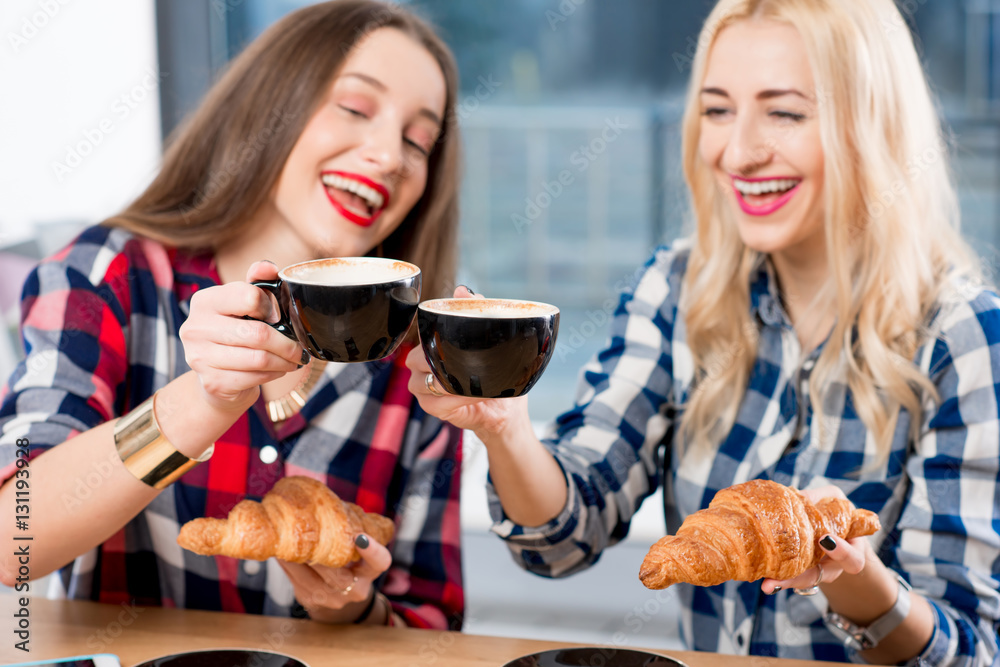 Young female friends dressed casually in checkered shirts sitting with coffee cups and croissants at