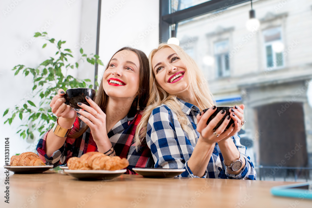 Young female friends dressed casually in checkered shirts sitting with coffee cups and croissants at