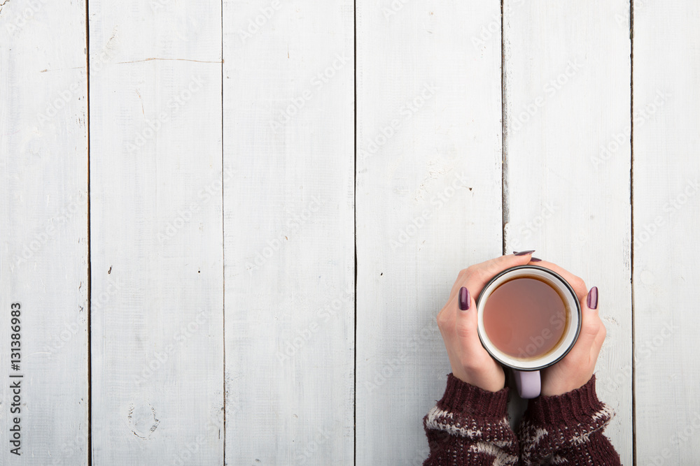 Womans hands in sweater holding cup of tea on the white wooden table