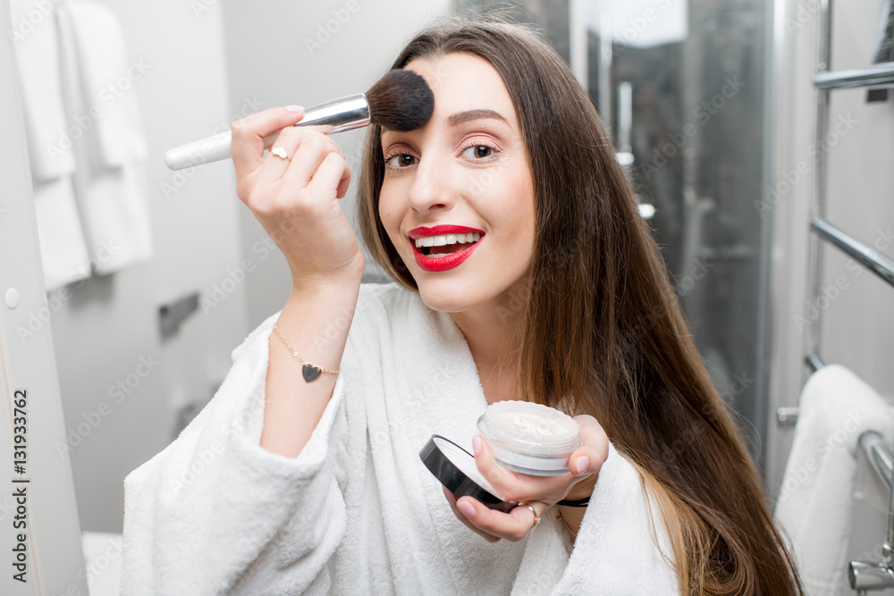 Young smiling woman in bathrobe applying powder in the bathroom