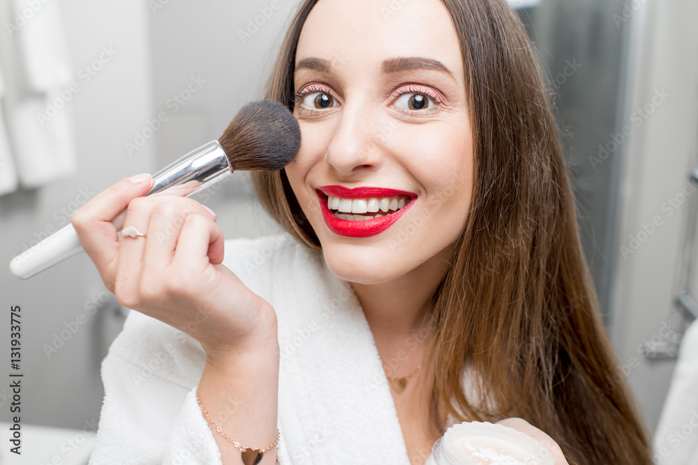 Young smiling woman in bathrobe applying powder in the bathroom