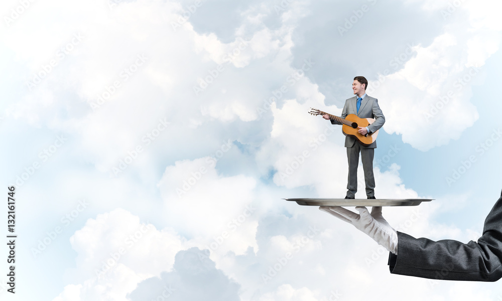 Businessman on metal tray playing acoustic guitar against blue sky background