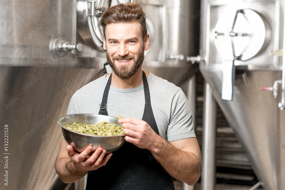 Portrait of a handsome brewer in uniform with hop in the bowl at the manufacturing with metal tanks