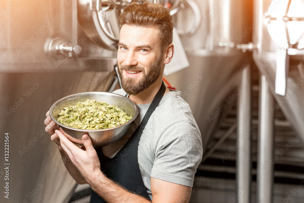 Portrait of a handsome brewer in uniform with hop in the bowl at the manufacturing with metal tanks