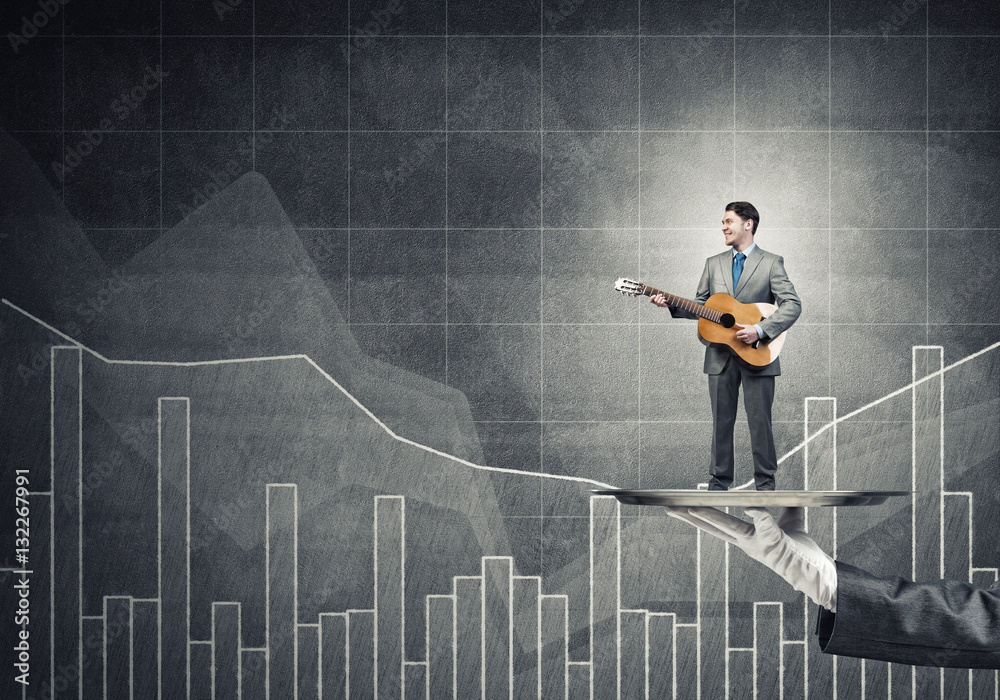 Businessman on metal tray playing acoustic guitar against concrete wall background with charts