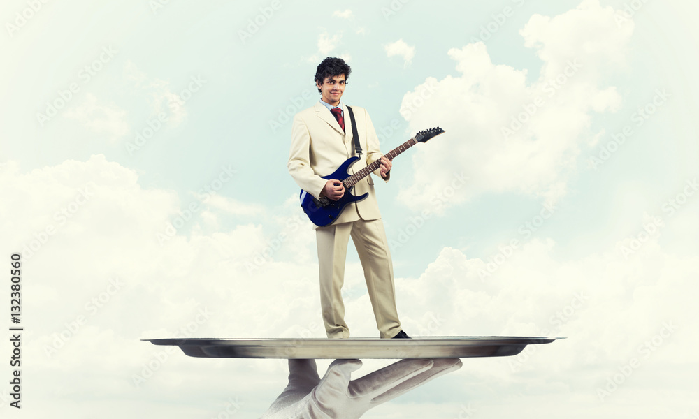 Businessman on metal tray playing electric guitar against blue sky background