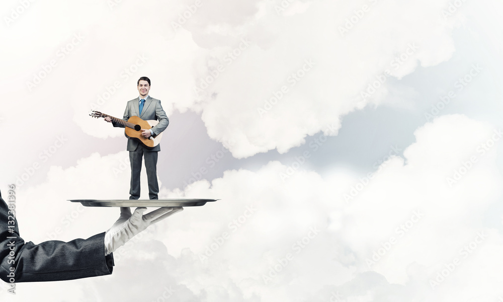 Businessman on metal tray playing acoustic guitar against blue sky background