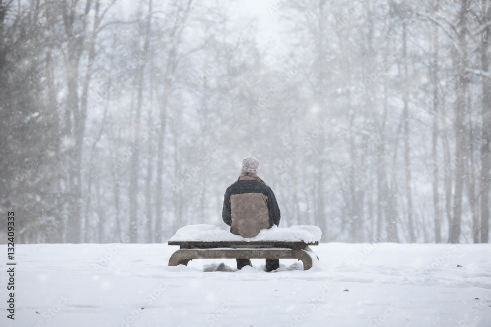 Lonely man sitting on the snowy benches in the park during the snowfall.