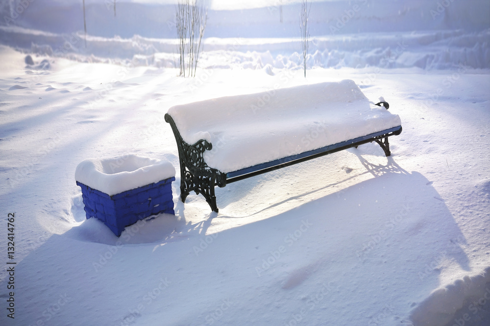 Bench in winter outdoors covered snow in a park town in the sunlight. Large snowdrifts.