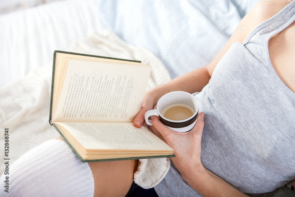 Girl reading book and drinking coffee in bed at home