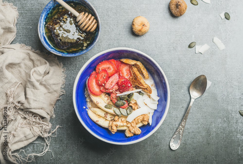 Yogurt, granola, seeds, fresh and dry fruits and honey in blue ceramic bowl over grey concrete backg