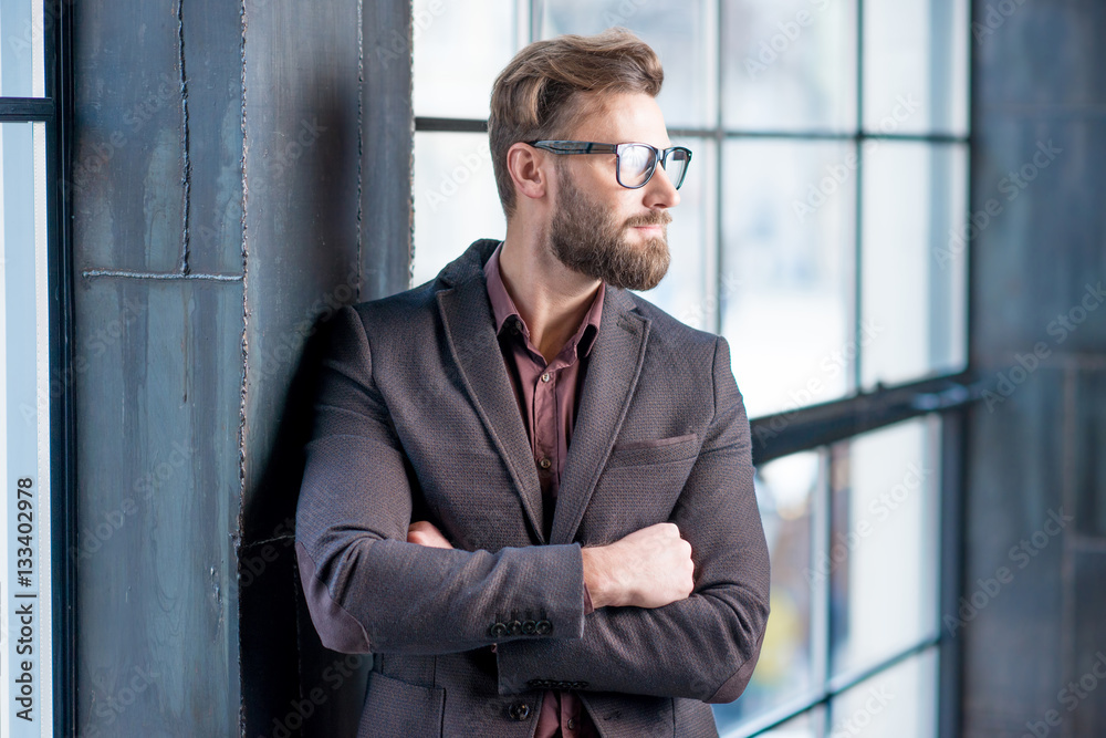 Portrait of caucasian bearded businessman dressed in suit with eyeglasses near window in the loft in