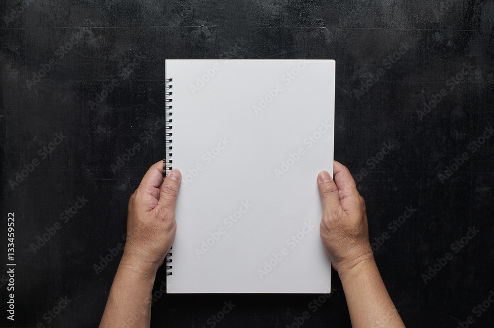 Wooden black office desk table with hand holding white blank notebook. top view