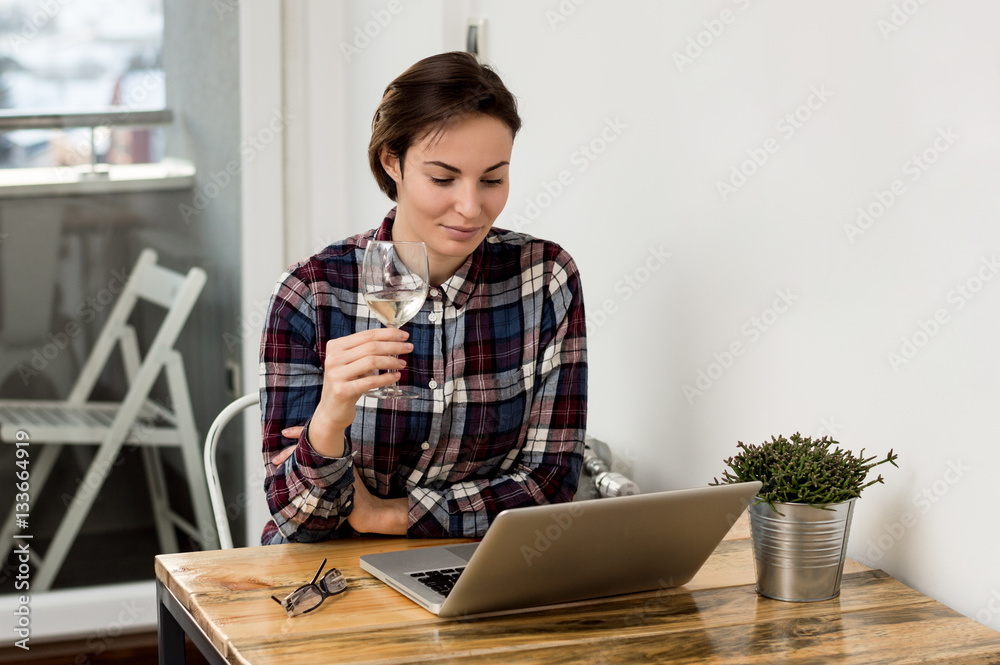 Pretty young brunette taking a sip of wine while using a compute