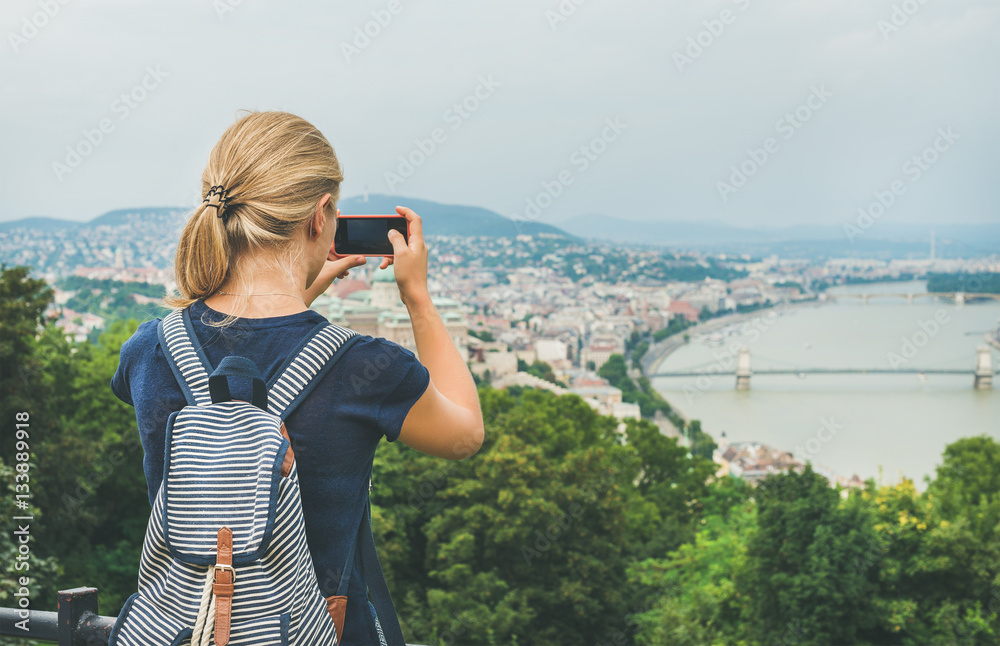 Young blond woman tourist making photoes of Danube river and Budapest from hill with smartphone, rea