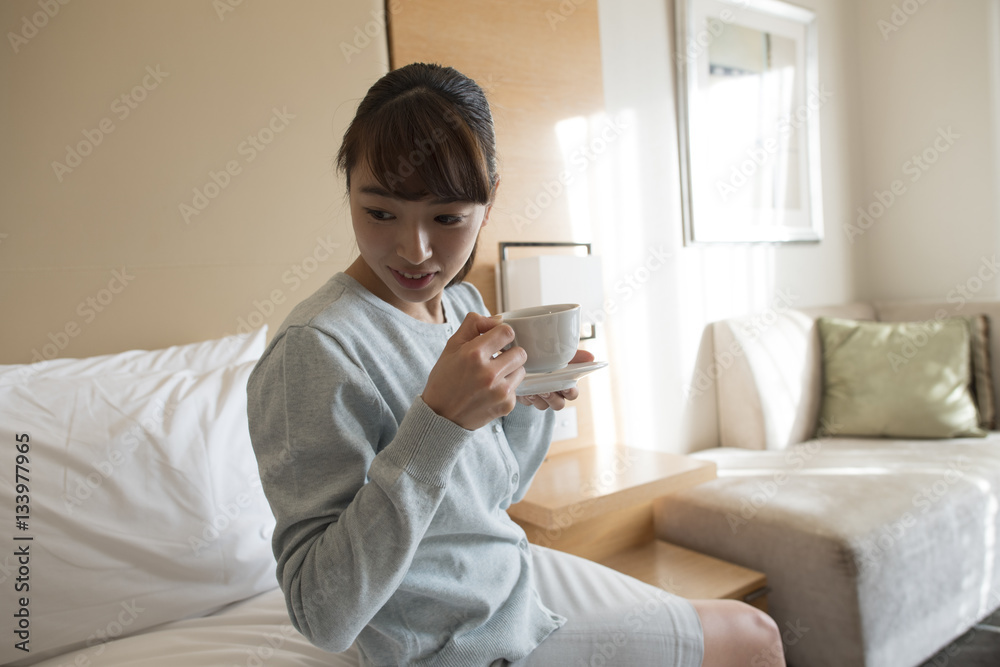 The woman is sitting in the bed in the hotel room and drinking coffee