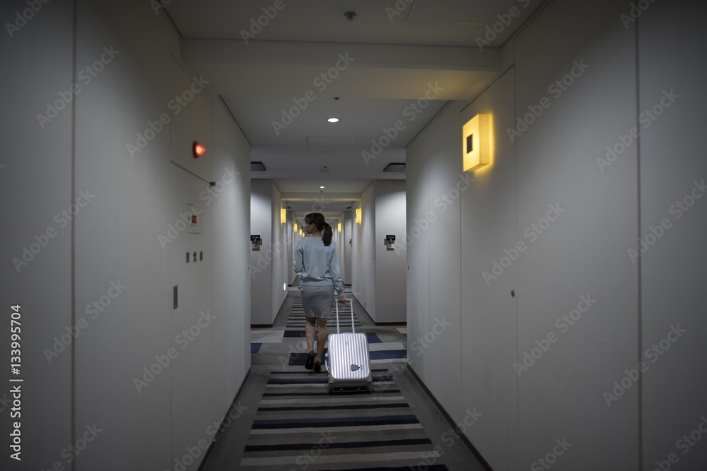 The woman is walking in the hotel corridor with a suitcase