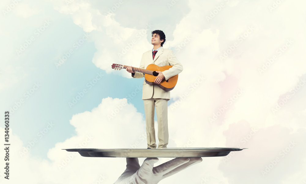 Businessman on metal tray playing acoustic guitar against blue sky background