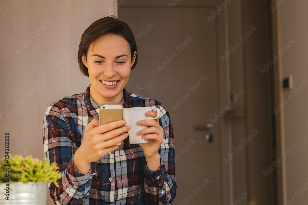 Happy beautiful girl using smart phone at home, drinking coffee