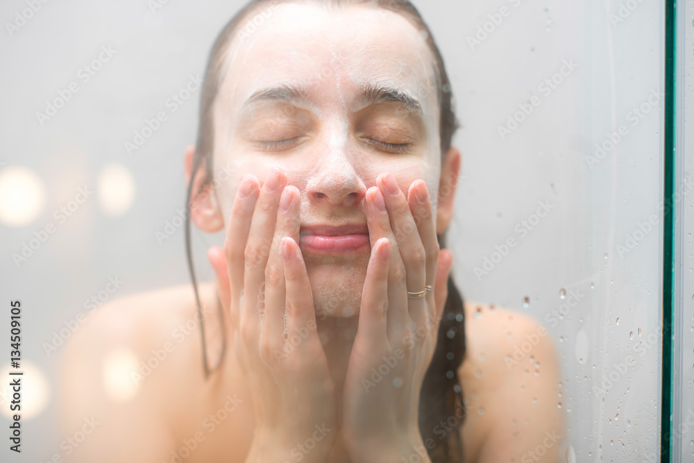 淋浴时，一个湿漉漉的脸上涂着肥皂的女人站在玻璃后面的特写肖像。Imag