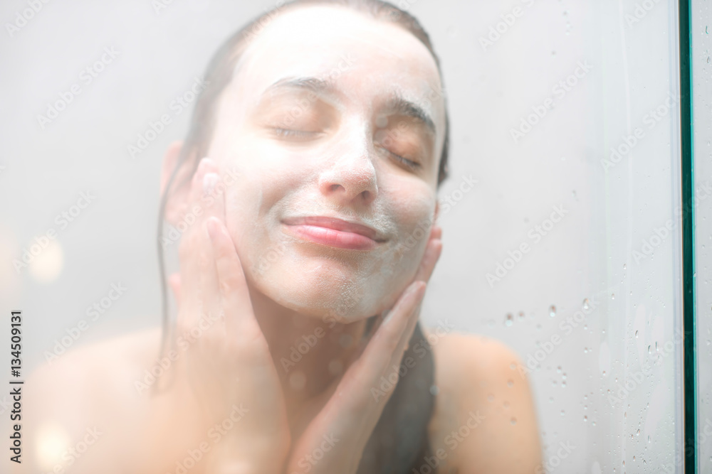 Close-up portrait of a woman with soap on her wet face standing behind the glass in the shower. Imag