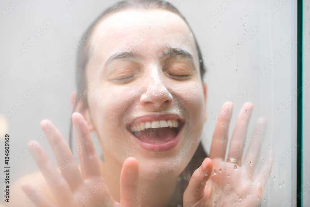 Close-up portrait of a woman with soap on her wet face having fun touching the glass in the shower. 