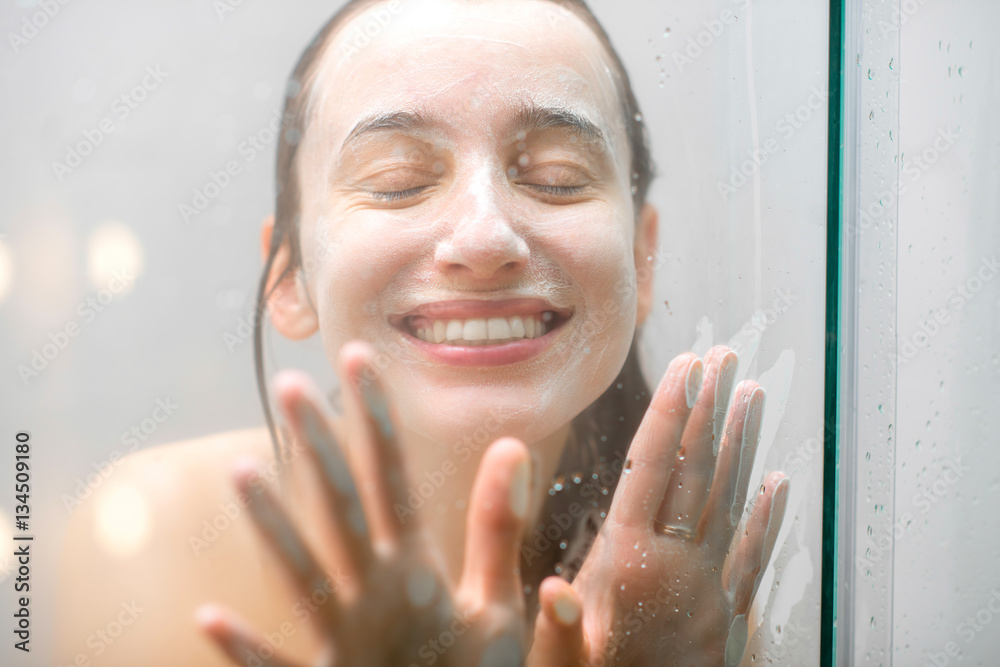 Close-up portrait of a woman with soap on her wet face having fun touching the glass in the shower. 