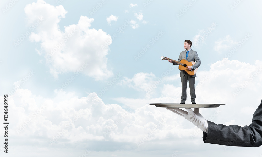 Businessman on metal tray playing acoustic guitar against blue sky background