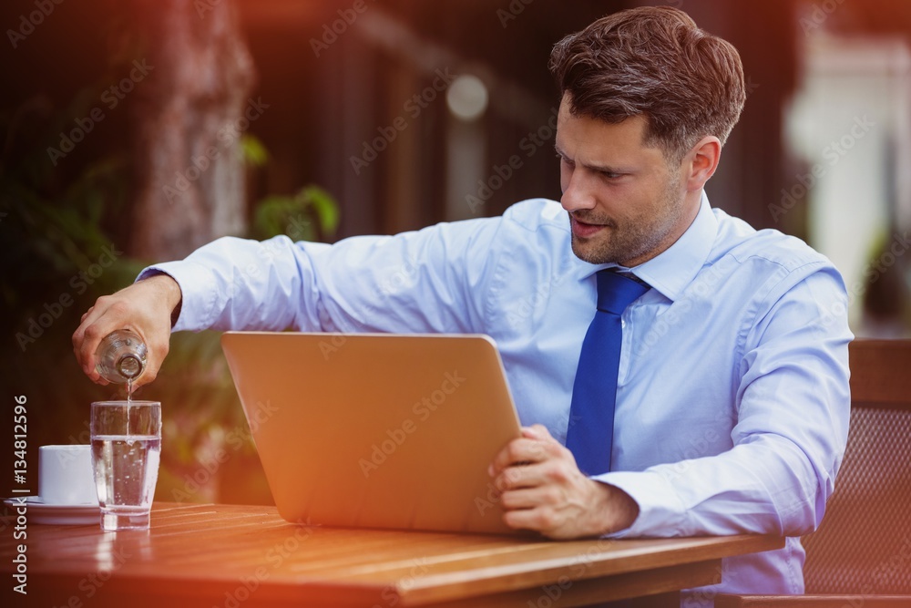 Handsome businessman pouring water in glass while using laptop