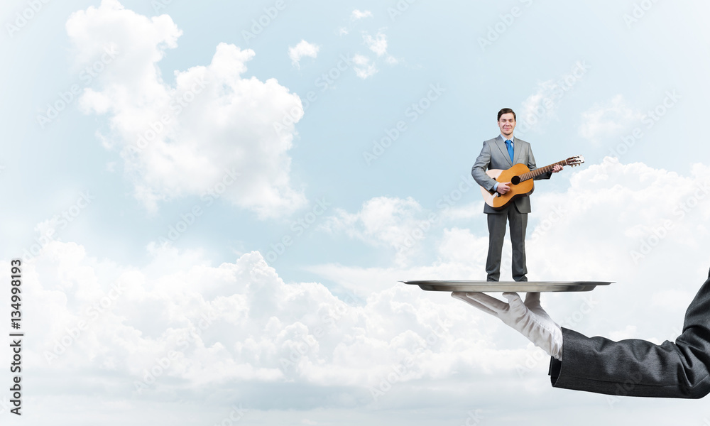 Businessman on metal tray playing acoustic guitar against blue sky background