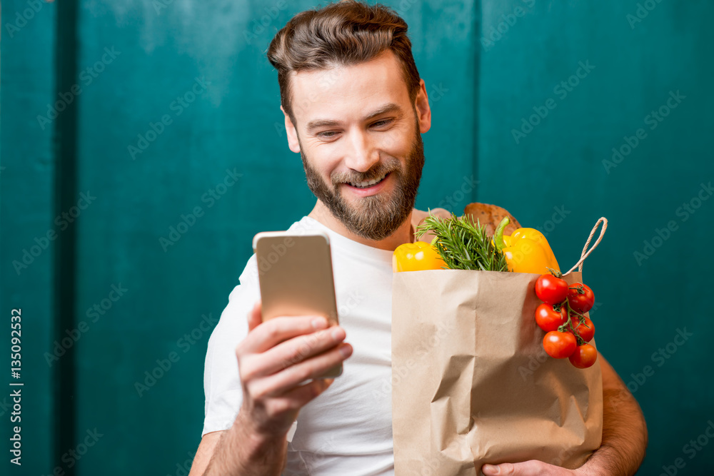 Man making online purchase with smart phone holding paper bag full of healthy food on the green back