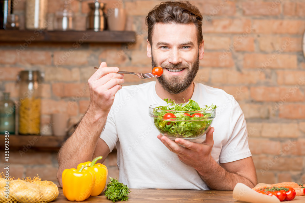 Handsome bearded man in white t-shirt eating salad with tomatoes in the kitchen. Healthy and vegan f