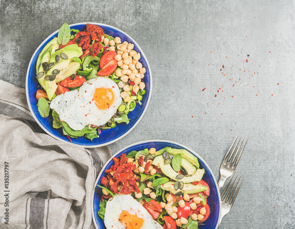 Healthy breakfast with fried egg, chickpea sprouts, seeds, vegetables and greens on bowls over grey 