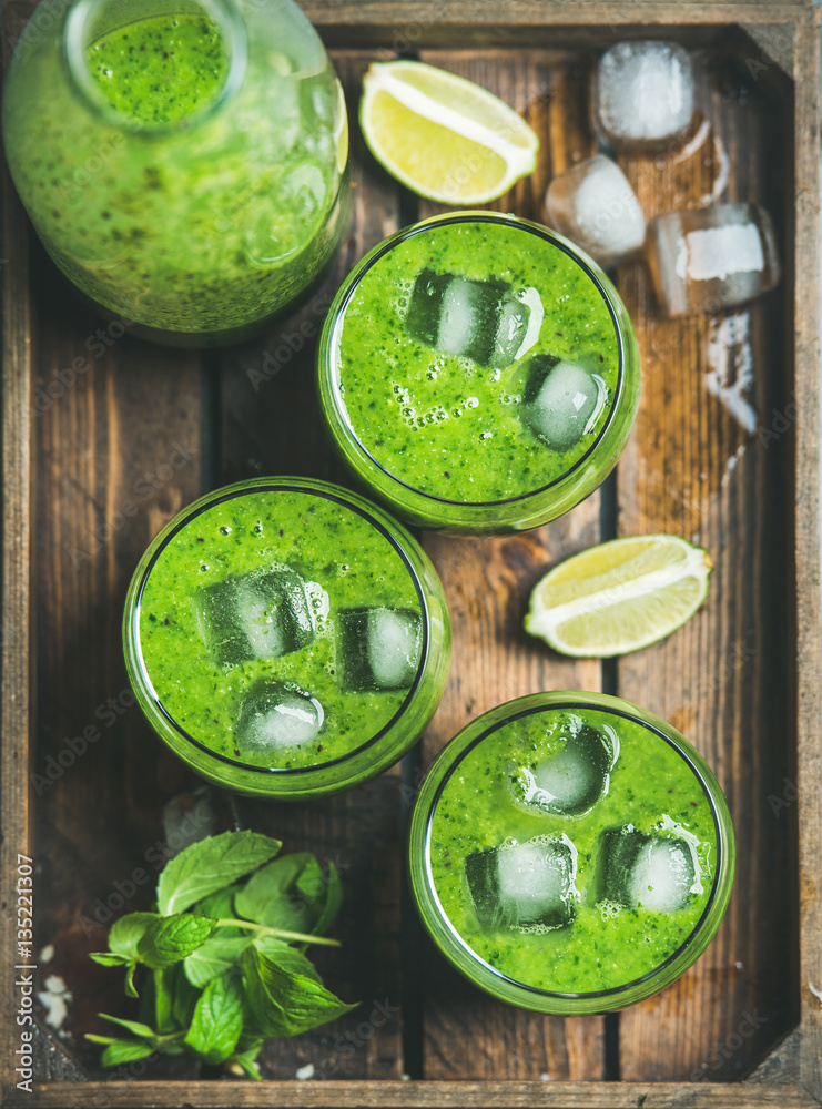 Fresh green smoothie in bottle and glasses with ice cubes, mint and lime in wooden tray, top view, s