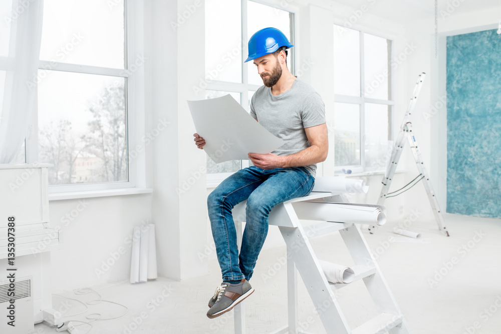 Handsome repairman or builder in helmet working with drawings on renovation of apartment interior