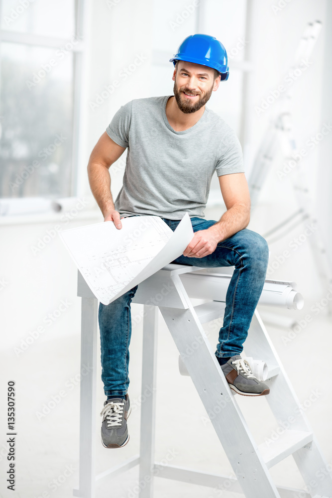 Portrait of a handsome builder, foreman or repairman in the helmet sitting with drawings on ladder i