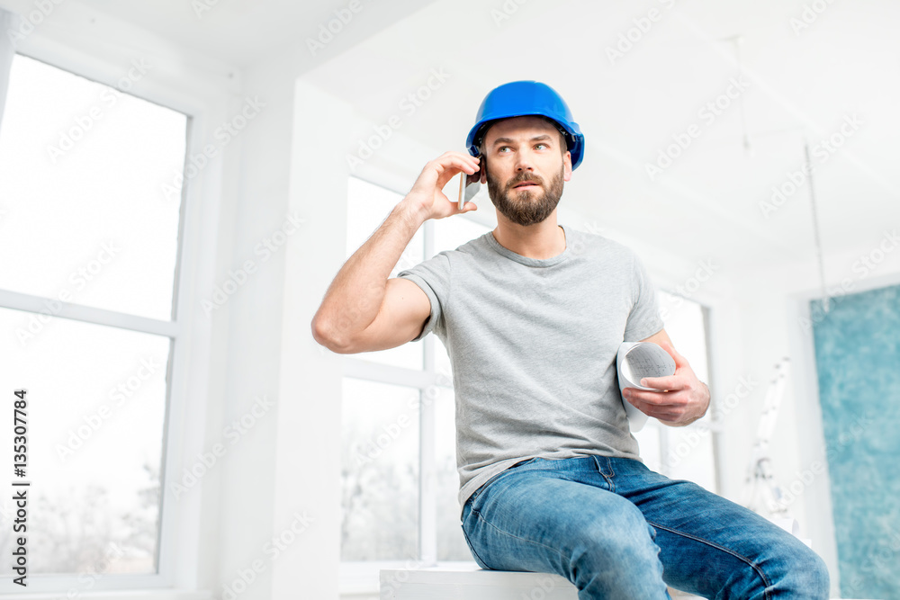 Handsome repairman or builder in helmet talking with phone with drawings in the white interior