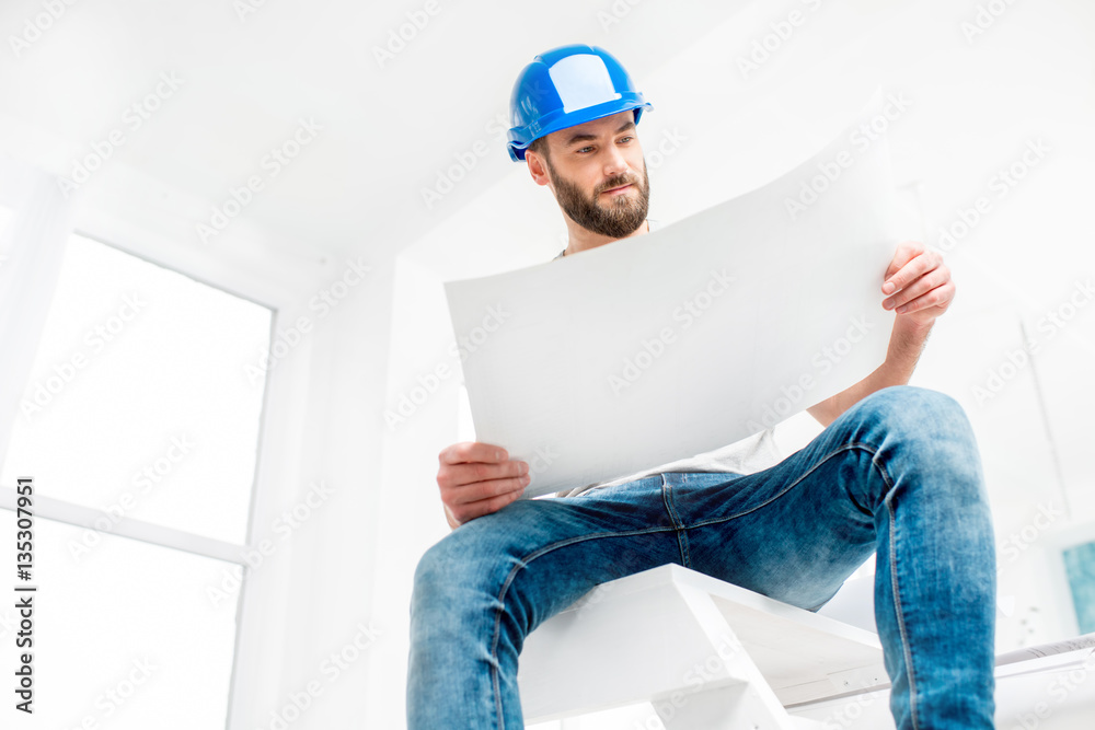 Portrait of a handsome builder, foreman or repairman in the helmet sitting with drawings on ladder i