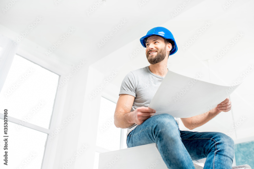 Portrait of a handsome builder, foreman or repairman in the helmet sitting with drawings on ladder i