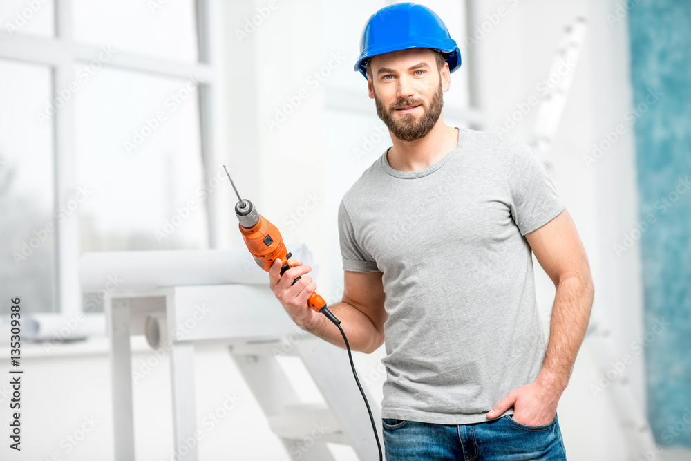 Portrait of a handsome repairman with drill and helmet in the bright interior