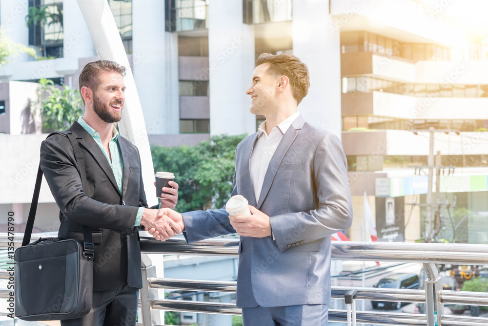 Two westerner businessman make hand shake on public walk way with cup of hot coffee in hand