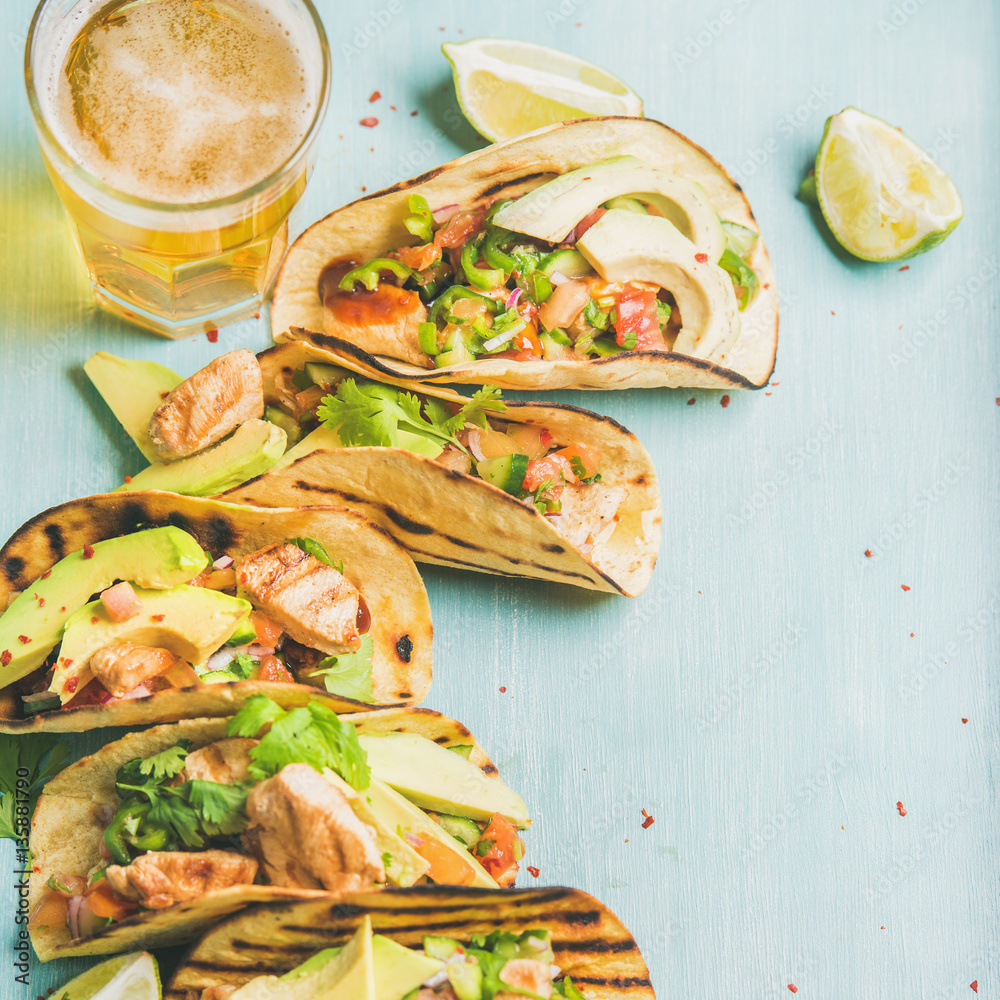 Healthy corn tortillas with grilled chicken, avocado, fresh salsa, limes and beer in glass over blue